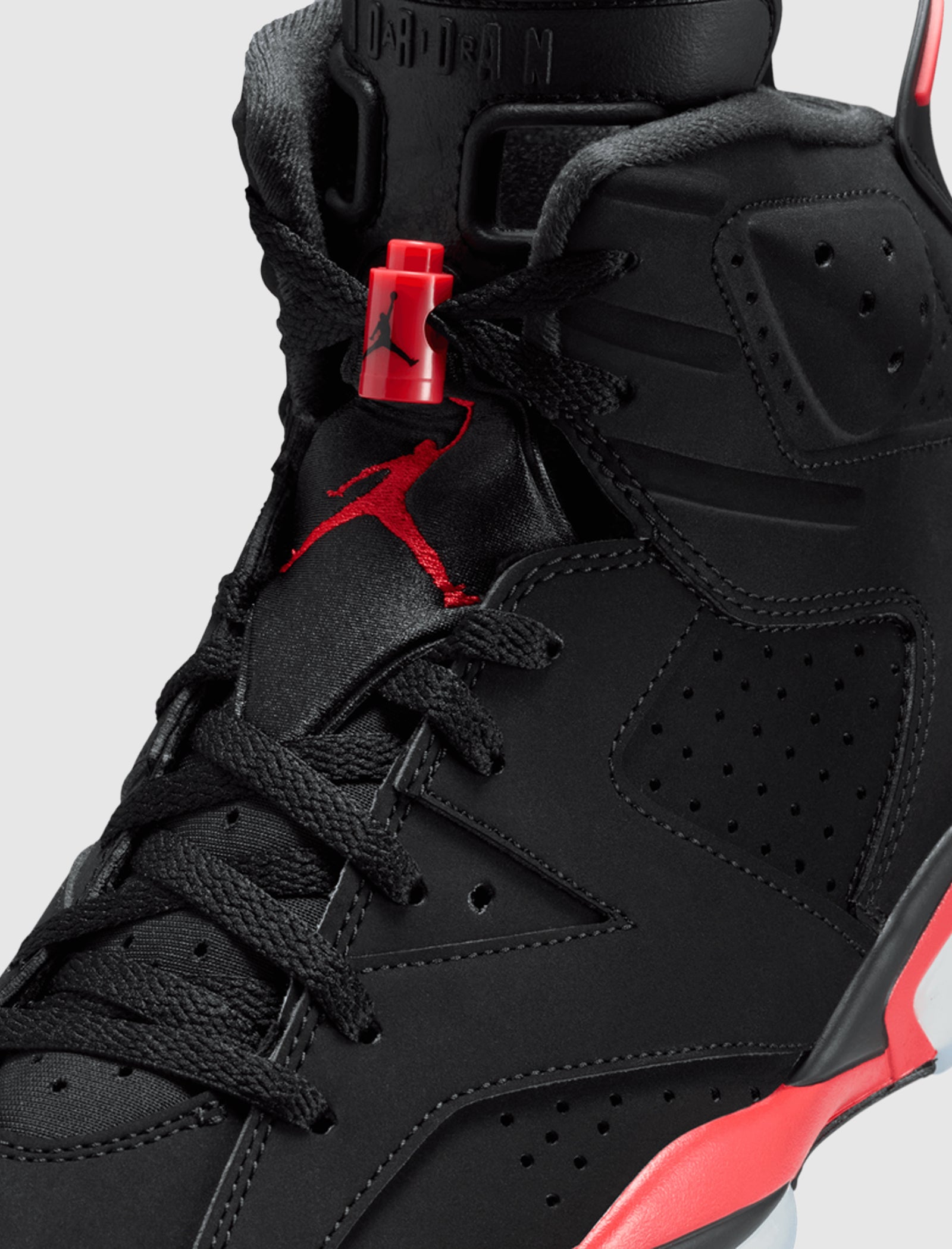 Close-up of the AIR JORDAN 6 RETRO "INFRARED SALESMAN" sneaker, featuring black perforated nubuck leather, black laces, red accents like a red Jumpman logo and lace lock, plus a cushioned foam midsole for comfort.