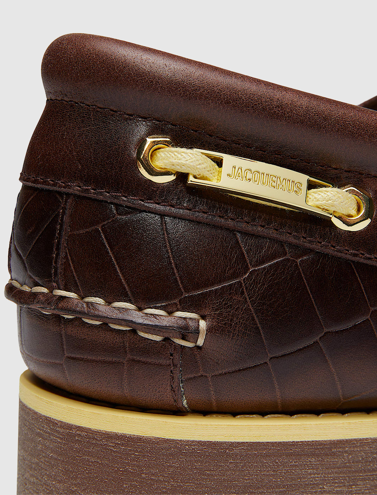 Close-up of a brown crocodile-embossed leather TIMBERLAND X JACQUEMUS 3 EYE BOAT SHOE with a cream-colored lace threaded through a gold-tone metal plate featuring the JACQUEMUS logo.