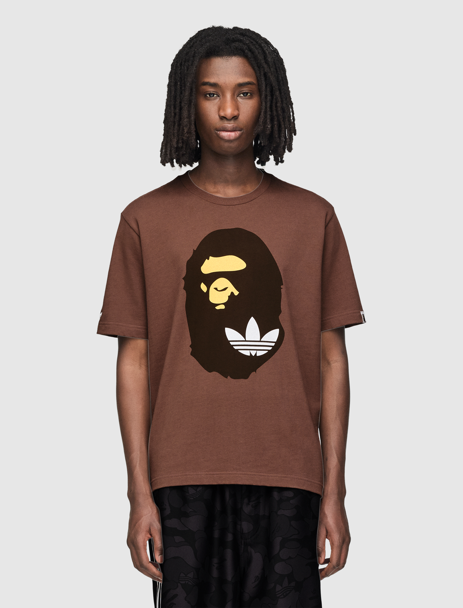 A person with medium-length dreadlocks wears the brown SSL TEE with a bold black and yellow graphic, paired with black patterned pants, standing against a plain light background.