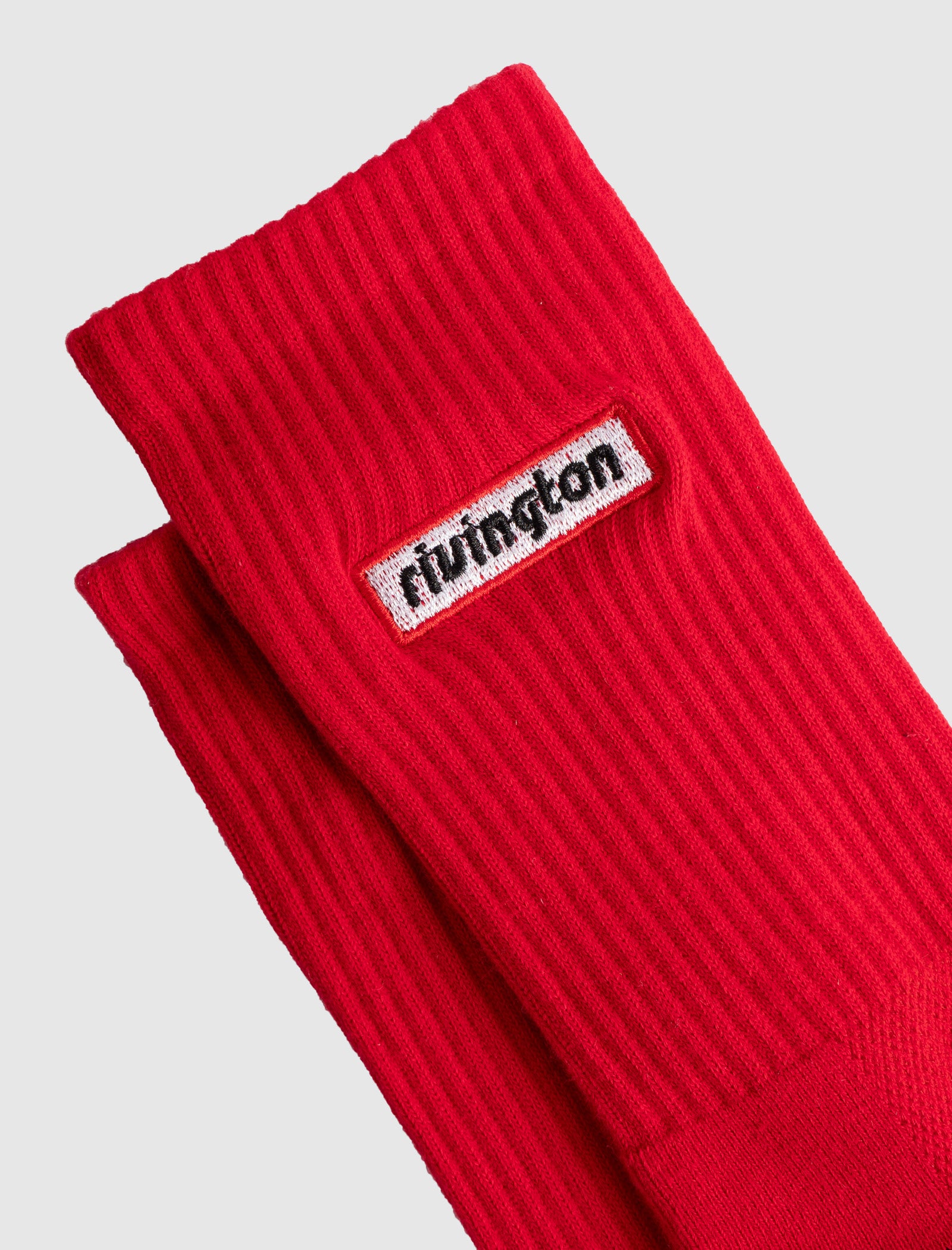 RIVINGTON SOCKS: Bright red ribbed crew socks with a rectangular white patch labeled "rivington" in black. Made of cotton, neatly folded on a light gray background.