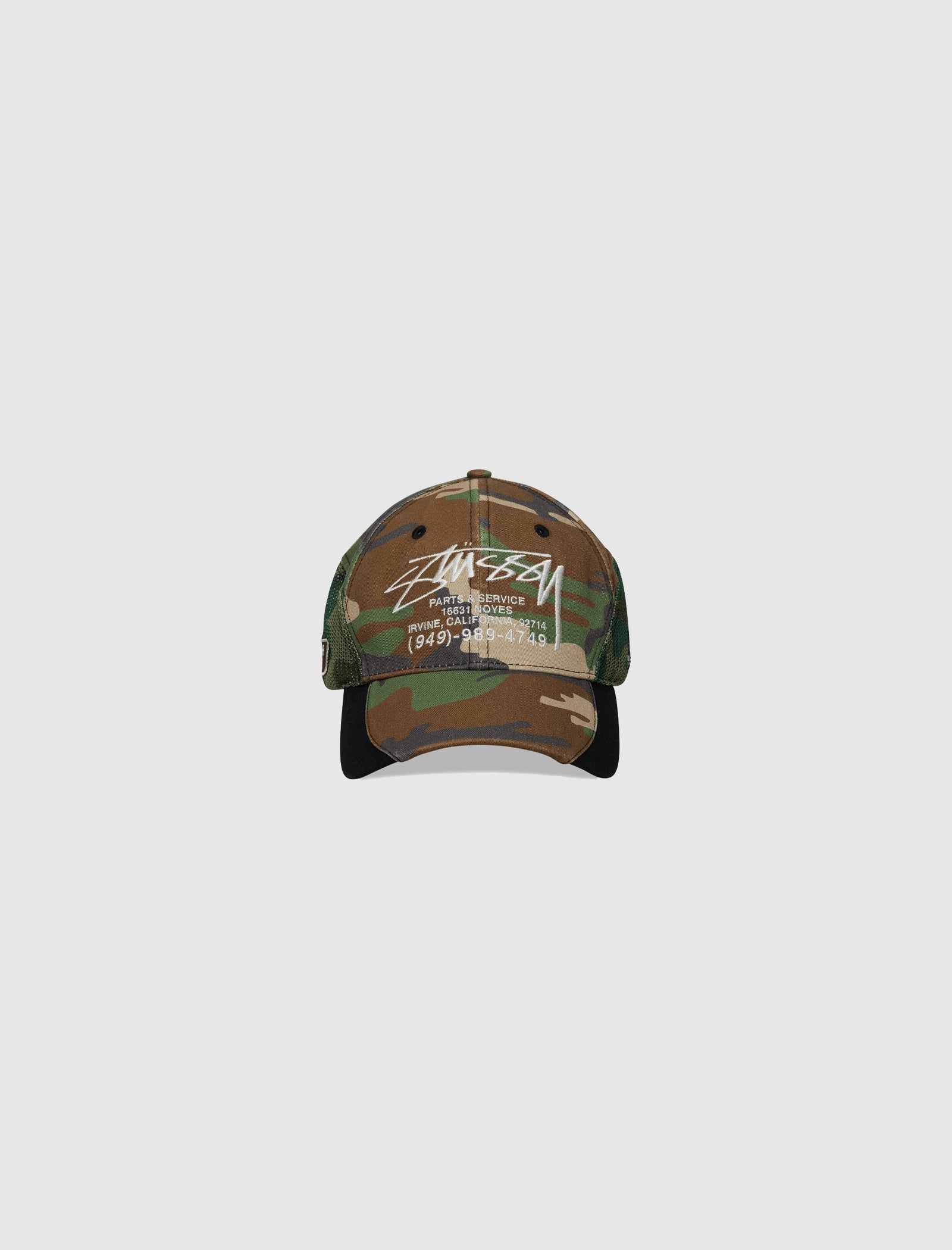 The LOW PROFILE SERVICE MESH STRAPBACK CAP features a camo print with white STÜSSY lettering, phone number, and INTERNATIONAL DESIGN CORP. on the front, a black brim, plain light gray background, and an adjustable snapback closure for a perfect fit.
