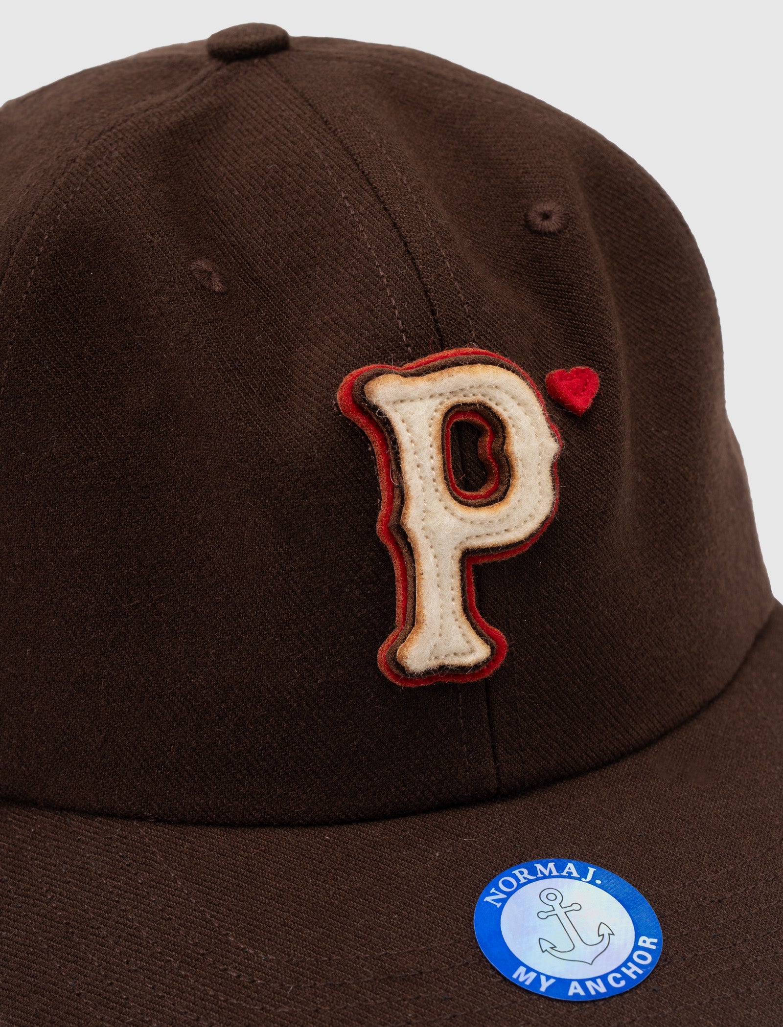 The WOOL BASEBALL "P" HAT is a brown wool snapback with a large textured "P," a small red heart above it, and a blue NORMAI MY ANCHOR sticker featuring an anchor. Adjustable for comfort.