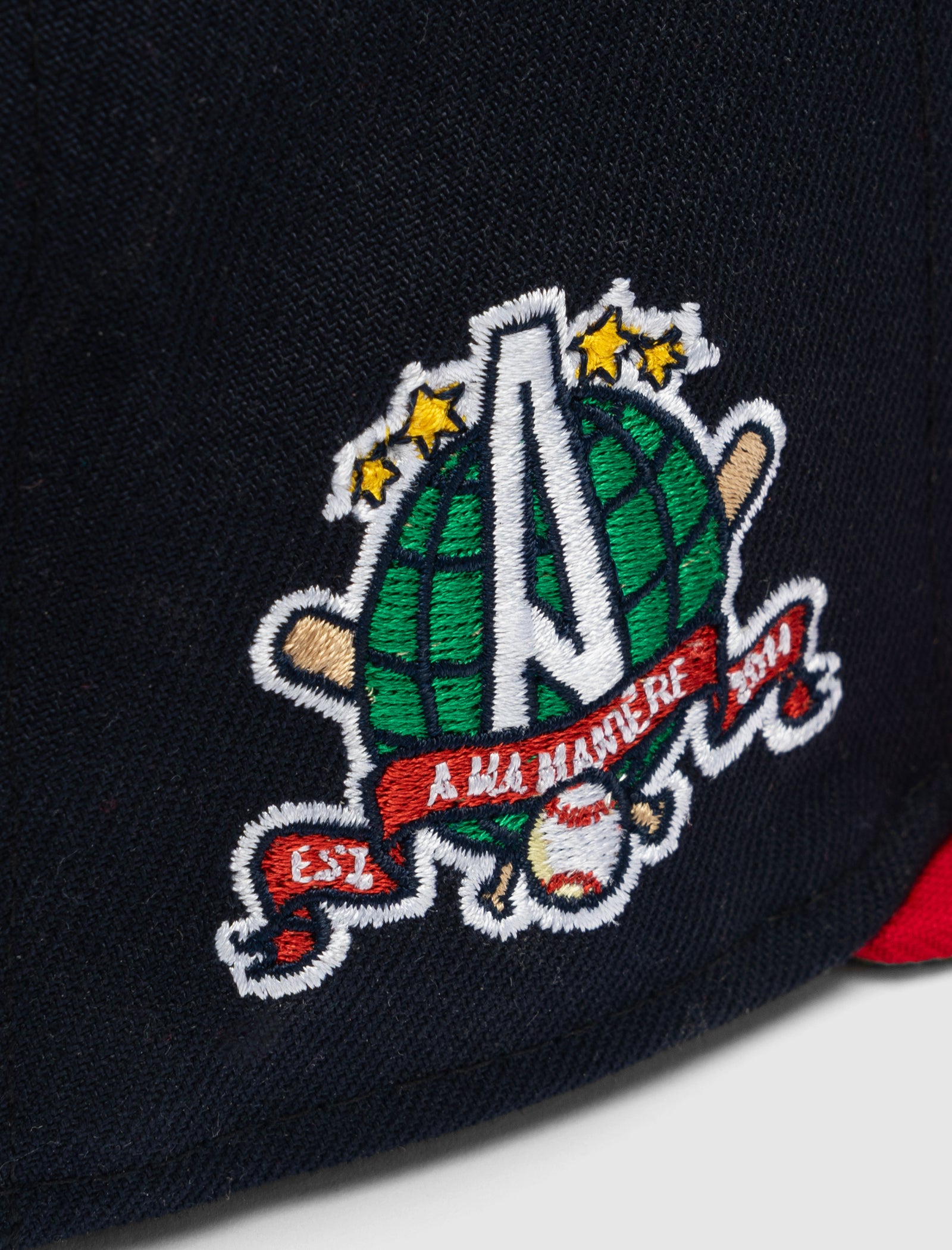 Close-up of the A MA MANIÉRE 5950 A LOGO HAT, featuring a navy fabric patch with a green globe, large white "A", three yellow stars, and a red banner reading "A Ma Maniere" and "EST. 2014" in white letters.