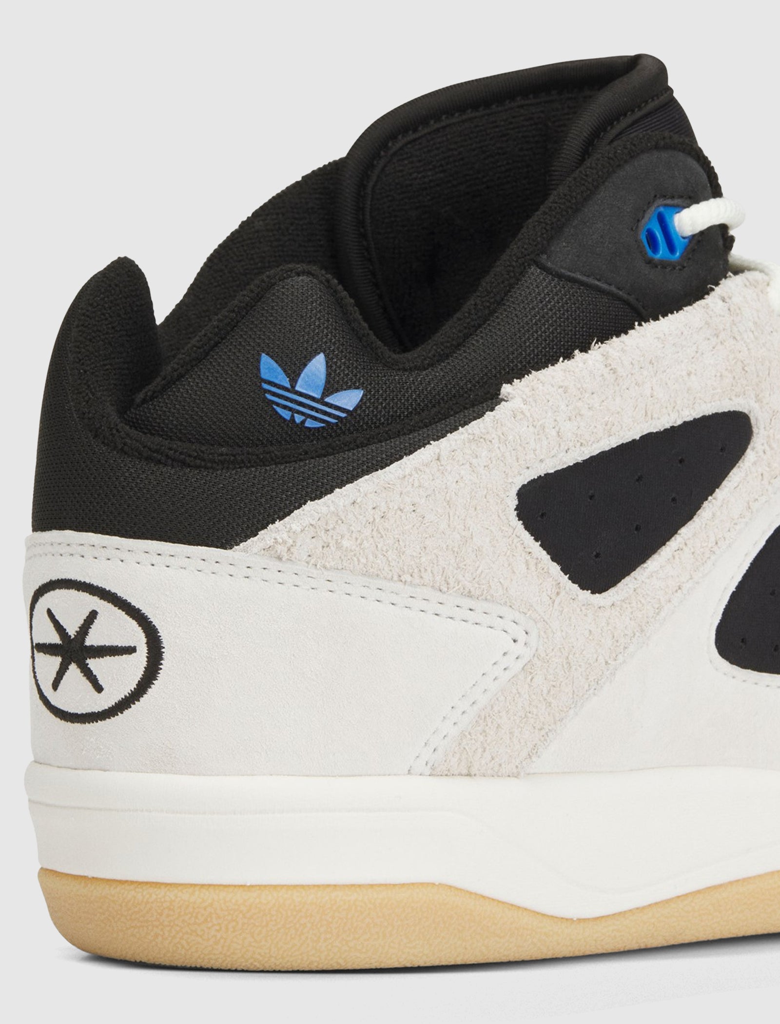 Close-up of the heel of the BADBO 1.0 "OFF WHITE/BLACK/GUM" mid top sneakers, featuring a black and white suede upper, blue Adidas Trefoil logo, circular star near the heel, white midsole, and tan rubber outsole.
