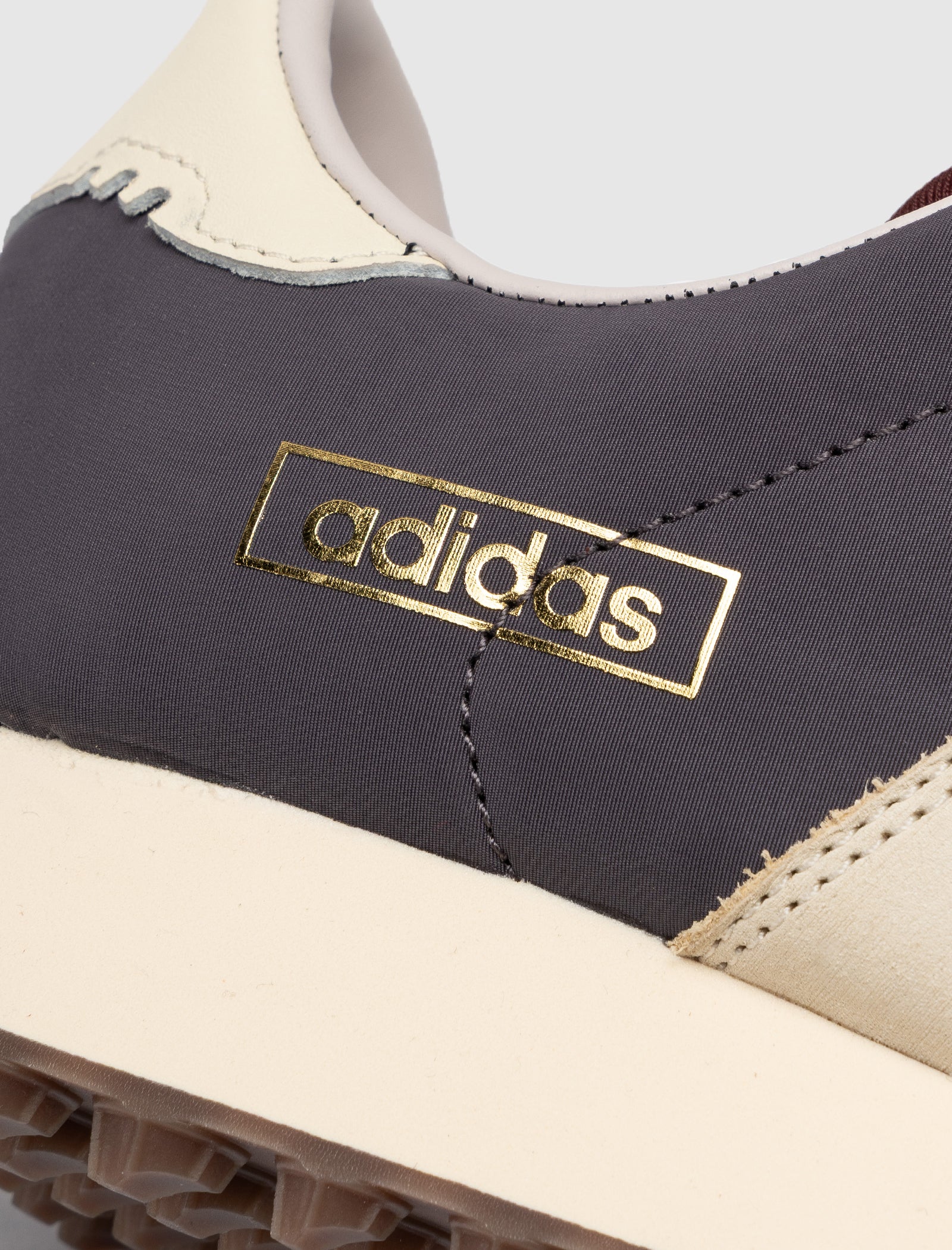 Close-up of the DND BW RUN HARLEM'S BEST sneaker, featuring a gold adidas logo on a dark gray fabric panel, with cream leather accents and a textured sole.