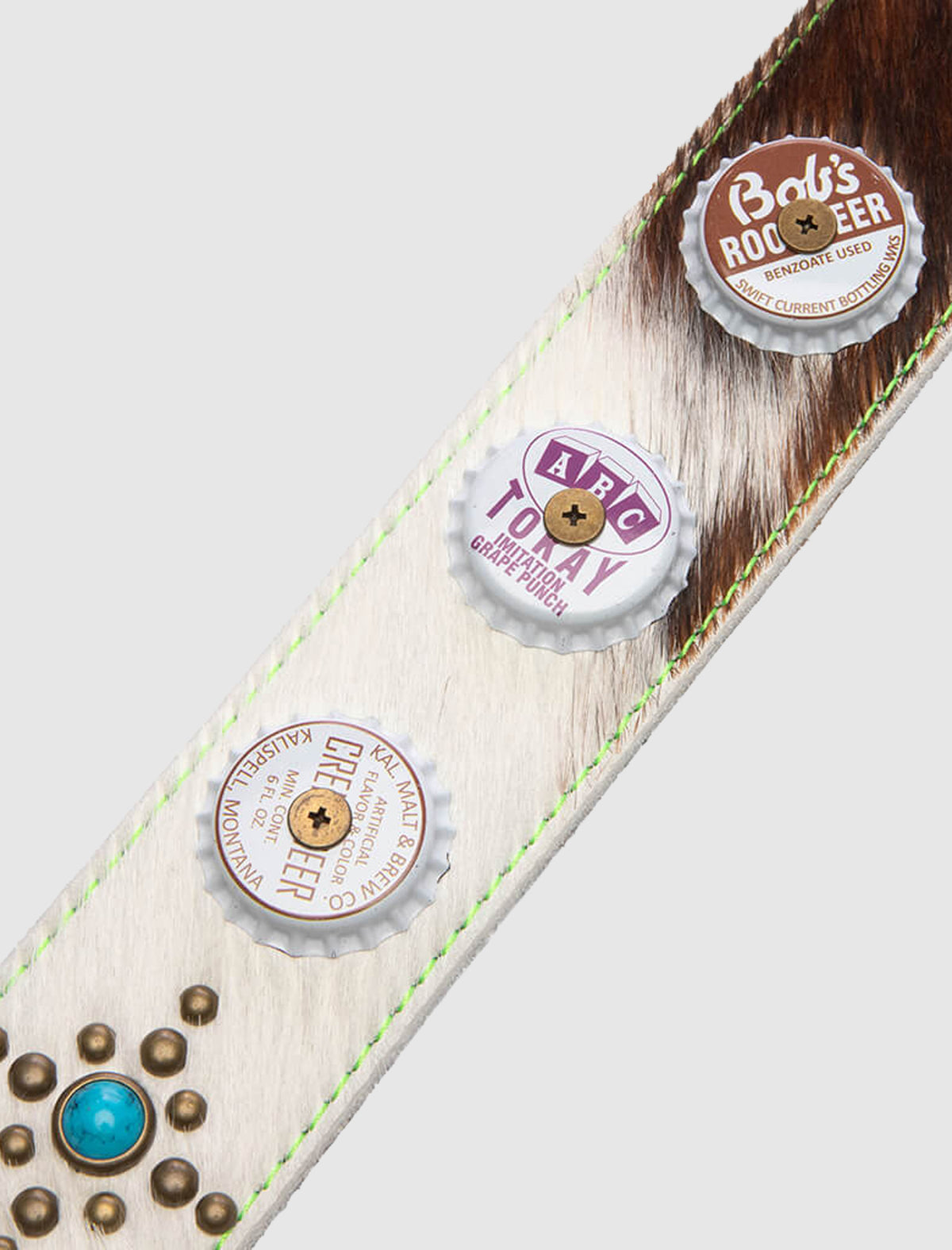 The CHASTITY BELT is a unique cowhide accessory belt accented with three metal bottle caps, each secured with a central brass rivet and finished with a turquoise stone surrounded by small metal studs.