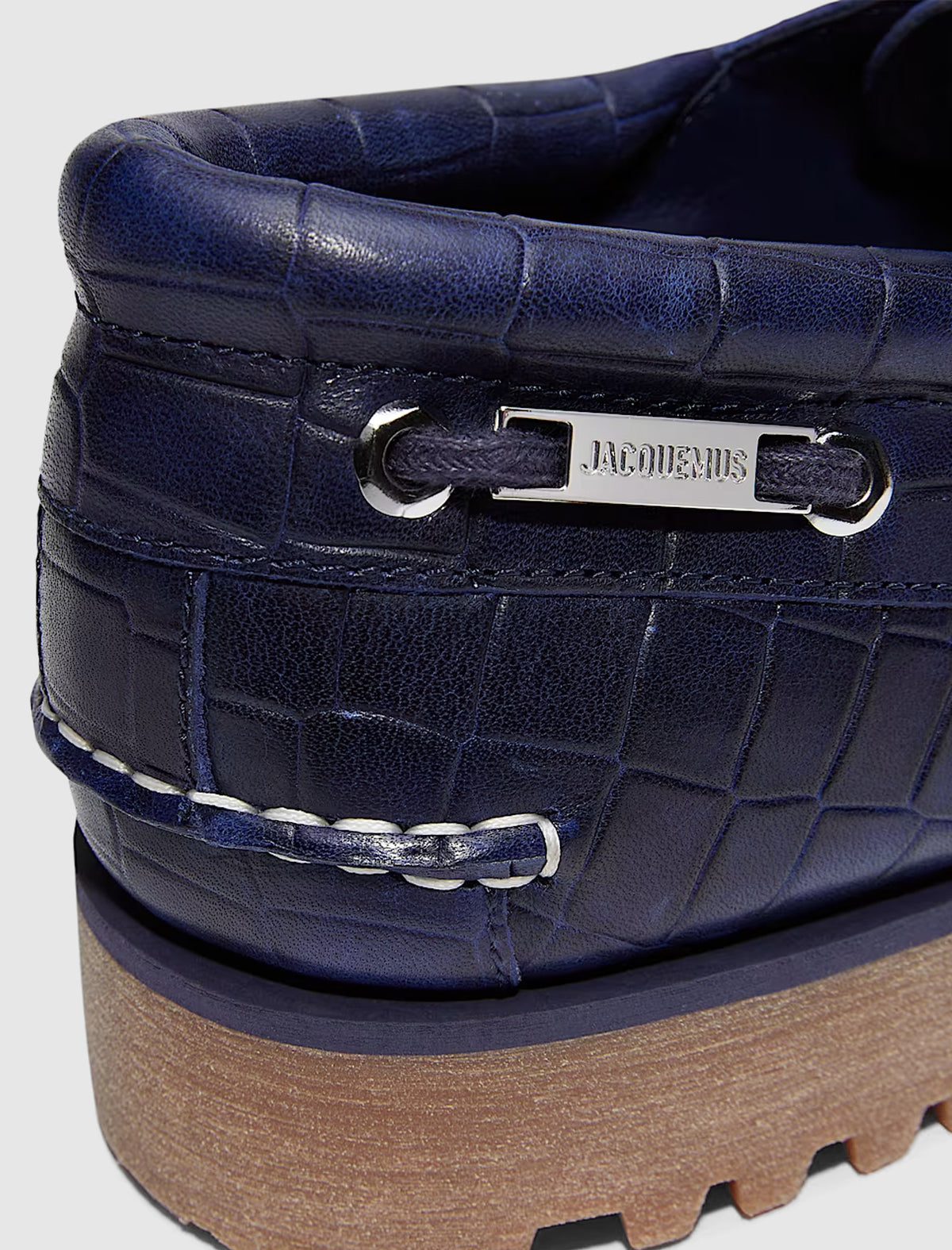 Close-up of the heel of a blue crocodile-embossed leather TIMBERLAND X JACQUEMUS 3 EYE BOAT SHOE, featuring a silver metal plaque engraved with the brand name.