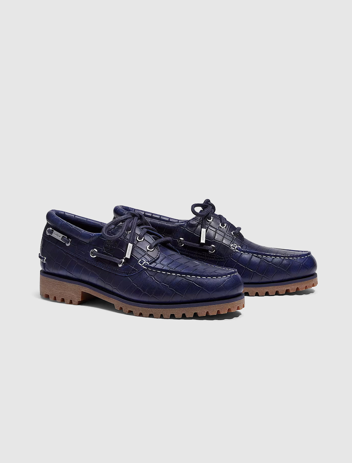 A pair of navy blue crocodile-embossed leather TIMBERLAND X JACQUEMUS 3 EYE BOAT SHOE featuring lace-up fronts and thick, serrated gum soles.
