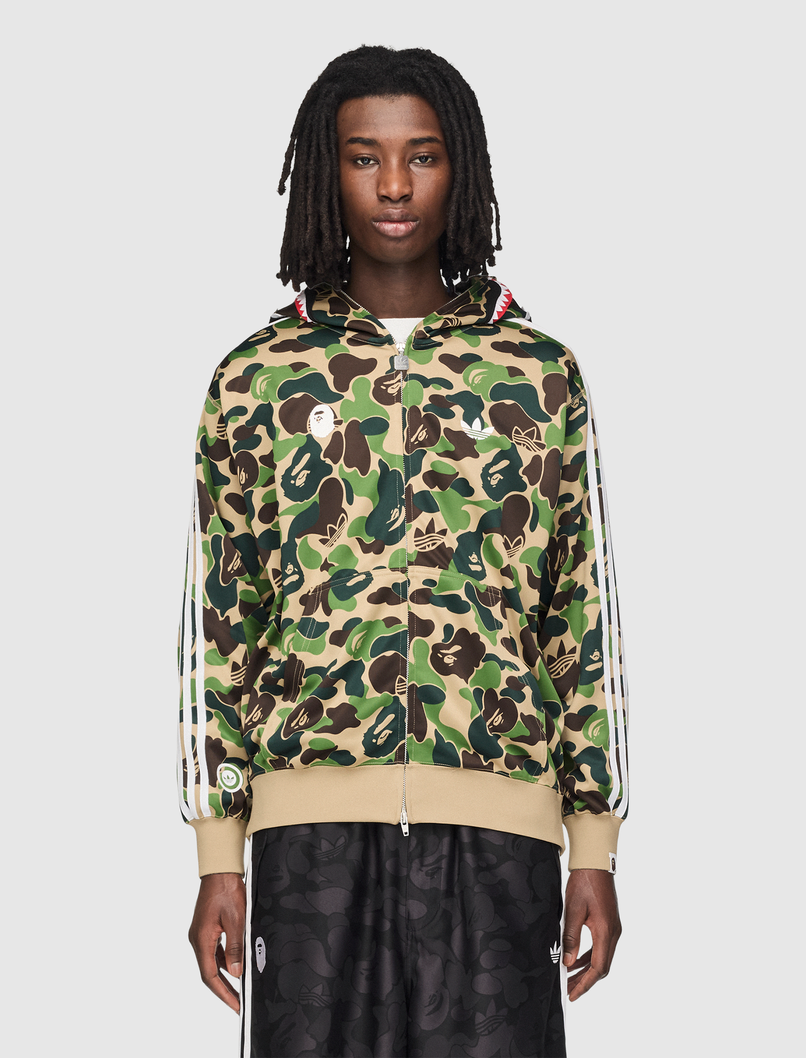 A person with long, dark dreadlocks wears the SHARK TRACK HOODIE in green camo with face graphics, paired with black patterned shorts, standing against a plain light gray background.