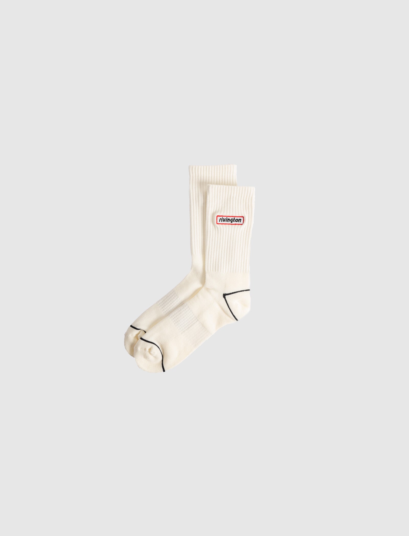 A pair of RIVINGTON SOCKS—white cotton crew socks with ribbed tops, black toe trim, and a red “thisisneverthat” logo patch on the sides—displayed against a plain light gray background.