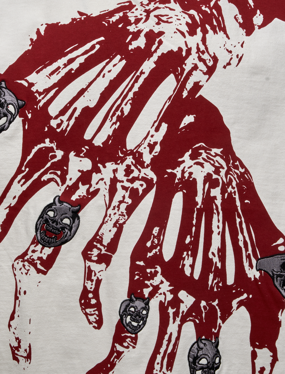 Close-up of a CROSS T SHIRT featuring a large, dark red graphic of a skeletal hand with small, embroidered grey devil masks placed on the finger bones.